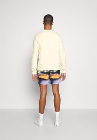 Felpa gialla in pile abbinata a pantaloni corti gradient multicolore con tonalità di arancione e viola, calze bianche e sneakers bianche.