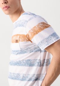 Camiseta de algodón blanca con rayas horizontales en azul y naranja, con un detalle de bolsillo y un corte relajado.