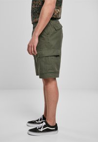 Pantaloni cargo verde oliva con multiple tasche, realizzati in tessuto leggero. Indossati con sneakers nere con lacci e dettagli bianchi.