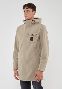 Giacca impermeabile beige con colletto alto, tasca frontale e una patch con logo. Presenta un design elegante e moderno e un tessuto testurizzato.