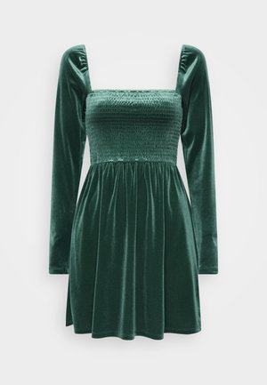 Robe en velours vert avec un corsage ajusté à smocks, décolleté carré et longues manches bouffantes ; jupe évasée avec des plis pour plus de volume.