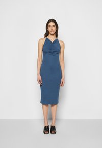Vestido de comprimento médio, sem mangas, em tecido azul escuro com textura franzida na área do busto, silhueta ajustada e barra arredondada. Usado com mules pretos.