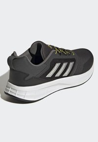 Zapatilla deportiva negra con parte superior de malla, cordones amarillos y negros, tres franjas plateadas y una suela blanca acolchada. Detalles texturizados en la parte superior.
