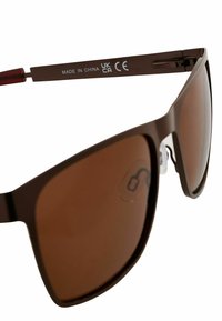 Braune Sonnenbrille mit dunkelbraunen, verspiegelten Gläsern. Kunststoffrahmen mit glatter Textur, ausgestattet mit durchsichtigen Nasenpolstern und gravierter Marke am Bügel.