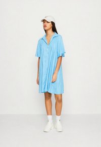 Mėlyna trumpomis rankovėmis marškinių suknelė su sagomis priekyje, minkšta tekstūra, laisvo kirpimo ir raukiniais. Dėvima su baltais sportbačiais ir kepure.