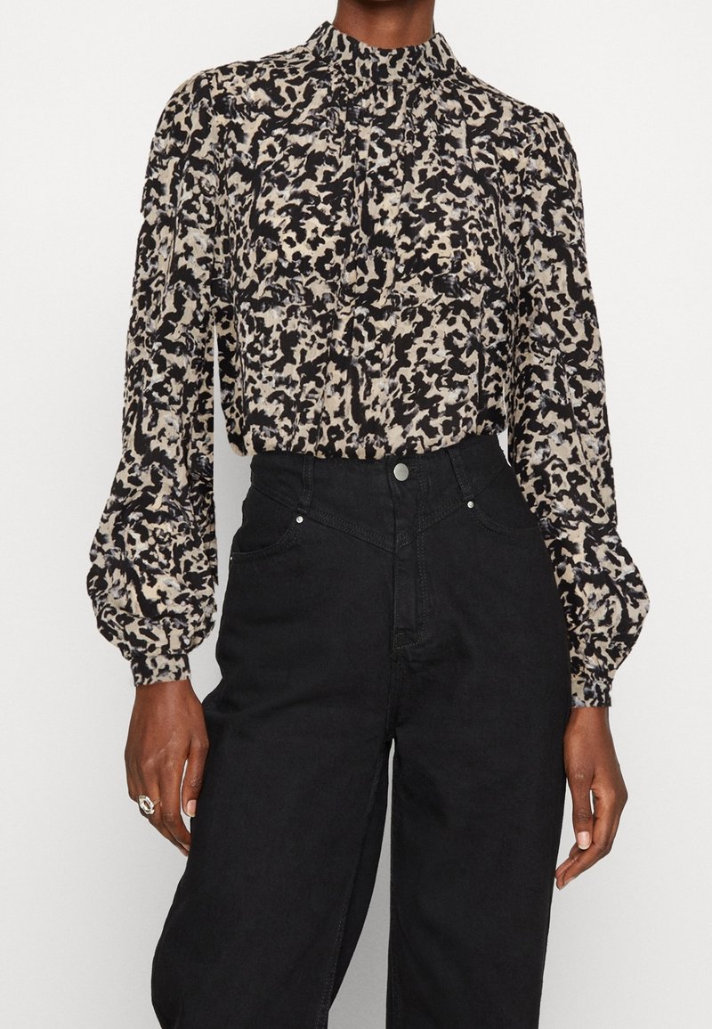 Femme portant un chemisier beige et noir à imprimé abstrait manches longues, rentré dans un pantalon noir taille haute avec boutons et poches.