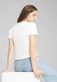 Camisa blanca de manga corta con un acabado texturizado, combinada con jeans de color azul claro que presentan un acento de cadena en el bolsillo trasero.