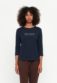 Vrouw met krullend haar, gekleed in een marineblauw "Tom Tailor" langarmshirt en een bruine broek, staand tegen een effen witte achtergrond.