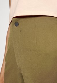 Pantalones verde oliva con una textura suave y un detalle de botón negro en la cintura, combinados con una parte superior de color rosa claro visible por encima.