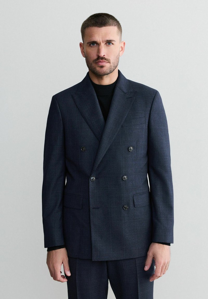 Blazer doppiopetto blu navy realizzato in tessuto testurizzato, con due tasche frontali e bottoni scuri, indossato sopra un dolcevita nero.