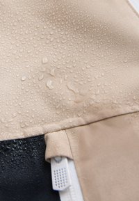 Gouttes d'eau sur un tissu beige de veste montrant un matériau résistant à l'eau et une partie du détail de la fermeture éclair blanche.