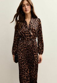 Klänning med leopardmönster och långa ärmar, tygband vid halsen, insydd midja och svävande kjol. Färgerna inkluderar svart, brunt och beige.