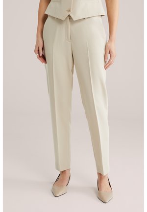 Naine kannab beeži värvi kohandatud pahkluumõõdulisi pükse, beeži värvi terava ninaga kontsakingi ja sobivat lühikest vestiga, käed kõrval.