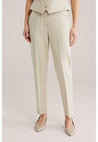 Vrouw draagt beige op maat gemaakte enkellange broek, beige gepunte hakken en een bijpassend cropped vest, met handen langs het lichaam.