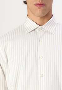 Camisa branca de botão com finas riscas beje, apresentando um colarinho clássico e cinco botões na parte da frente, feita de um tecido suave.
