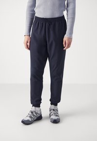 Osoba oblečená v proužkovaném světle šedém tričku s dlouhým rukávem, černých teplácích na šňůrku, bílých ponožkách a bílých-šedých turistických botách na bílém pozadí.