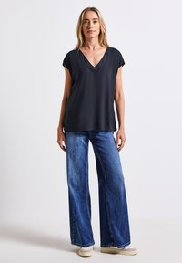 Blusa nera a V con maniche corte, abbinata a jeans in denim blu a gamba larga e sneakers beige. Texture liscia, vestibilità casual, design minimale.