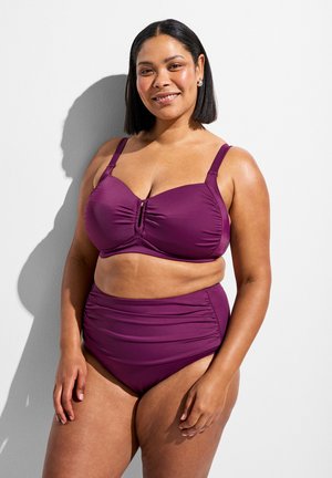 Naeratav naine õlani pikkade juustega, kellel on seljas lilla kaksosaline ujumisriietus valge seina ja varjuga taustal.