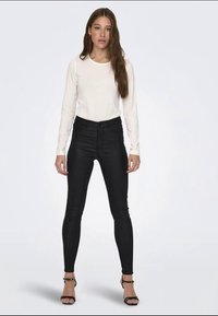 Black skinny jeans with a high waist, paired with a fitted white long-sleeve shirt. Model wearing strappy black heels, standing straight.