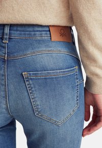 Los vaqueros de mezclilla azul presentan una superficie texturizada, costuras visibles y una etiqueta de cuero marrón con un logo en la cinturilla.