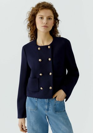 Femme aux cheveux roux bouclés portant une veste croisée bleu marine avec des boutons dorés et un jean bleu clair, debout devant un fond uni.