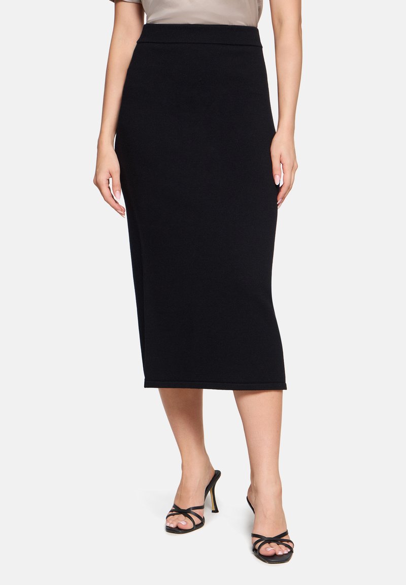 Femme portant une jupe midi noire ajustée et des sandales à talons hauts noires, debout devant un fond uni.