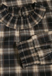 Black and beige checked fabric with a gathered ruffled neckline and elasticated cuffs, featuring a smooth texture and contrasting patterns.