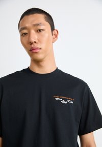 Black cotton t-shirt with a crew neckline, featuring orange embroidered text and three fish illustrations on the left chest.