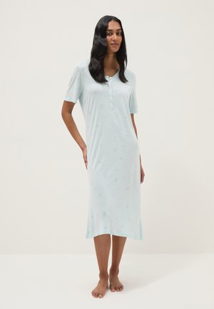 Donna in piedi a piedi nudi che indossa una camicia da notte azzurro chiaro al ginocchio con maniche corte e bottoni sul petto.