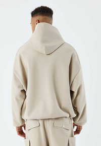Felpa beige oversize con un grande cappuccio strutturato. Il tessuto appare morbido e testurizzato, presenta spalle cadenti e una vestibilità rilassata.