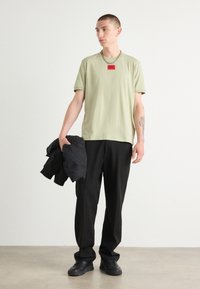 Tricou verde deschis cu un patch roșu pe piept, asortat cu pantaloni negri. Subiectul ține o jachetă neagră și poartă adidași negri.