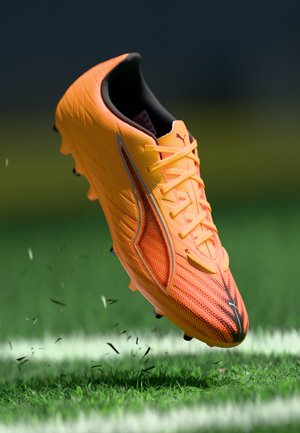 Scarpetta da calcio arancione con tomaia testurizzata, dettagli neri e tacchetti visibili, che si libra sopra l'erba con fili sparsi.