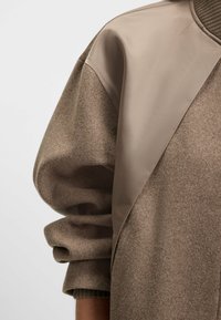 Ruskea villa-sekoiteswetteri, jossa on sileä, beige satiinipaneli olkapäissä. Sweaterissä on ribbikuvioinen hihansuu ja pehmeä, tekstuuriainen kangas.