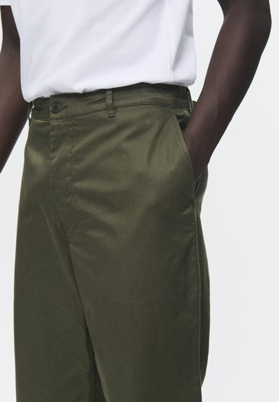 Olivgrüne Hose aus glattem Stoff, mit einem Knopfverschluss, seitlichen Taschen und einer taillierten Passform, kombiniert mit einem weißen Hemd.