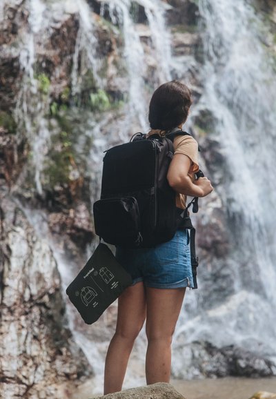 Zaino nero con zip e più scomparti. Il modello tiene una borsa nera piegata con illustrazioni di hardware bianco. Cascata sullo sfondo.