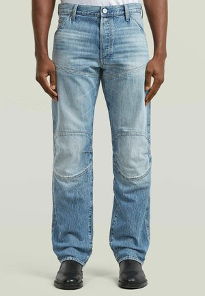 Uomo che indossa jeans di jeans azzurri con toppe sulle ginocchia, cuciture visibili e scarpe nere su uno sfondo semplice.