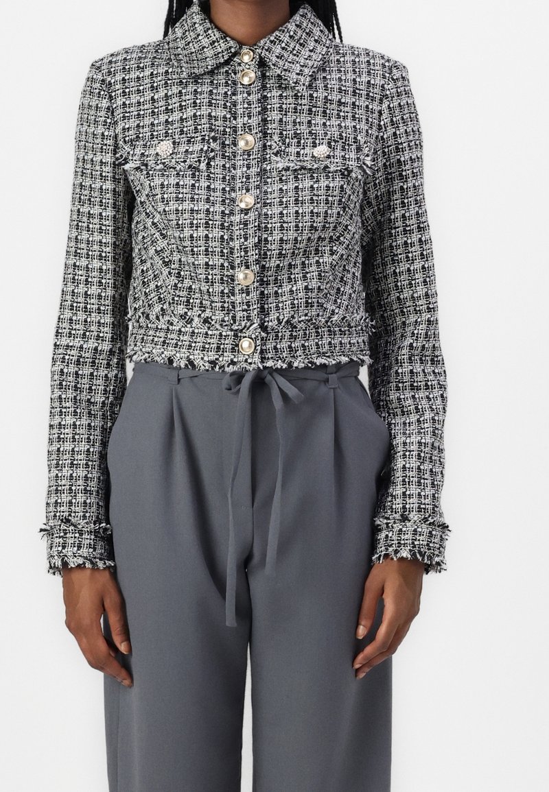 Femme portant une veste courte texturée noire et blanche avec des boutons en perle et un pantalon gris taille haute noué à la taille.