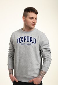 Grå sweatshirt med ribbad mudd och nederkant. Har "OXFORD UNIVERSITY" i fet marinblå skrift över bröstet. Mjuk tygtextur.