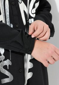 Zwarte shirt met grote zilveren print met druppels. Close-up van een dichtgeknoopte manchette met een gladde textuur en zwarte knoopdetails.