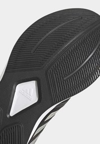 Suela de zapatilla negra con goma texturizada, patrones de relieve pronunciados y un logo blanco de Adidas en un acento en forma de diamante.