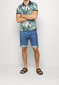 Koszulka polo z krótkim rękawem w tropikalny wzór liści i kwiatów, z ciemnym kołnierzem, w połączeniu z niebieskimi krótkimi dżinsami i czarnymi sandałami.