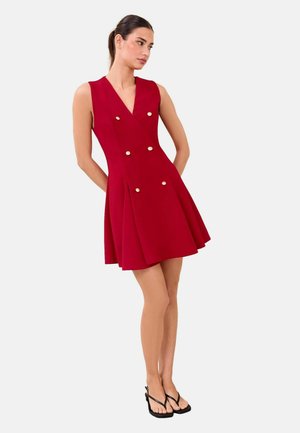 Kvinde iført en ærmeløs rød V-hals kjole med guldknapper og sorte højhælede sandaler, stående med hænderne bag ryggen.