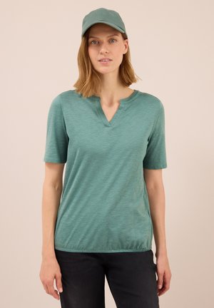 Femme portant une casquette verte, une chemise à manches courtes verte avec un col en V, et un pantalon noir, se tenant devant un fond beige uni.