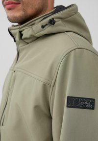 Veste vert olive claire avec une texture lisse, dotée d'une capuche à cordon de serrage, de accents noirs et d'un écusson de logo sur la manche.