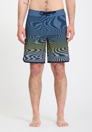 Hombre con pantalones cortos de baño con patrón ondulado azul y amarillo, de pie descalzo contra un fondo blanco liso.