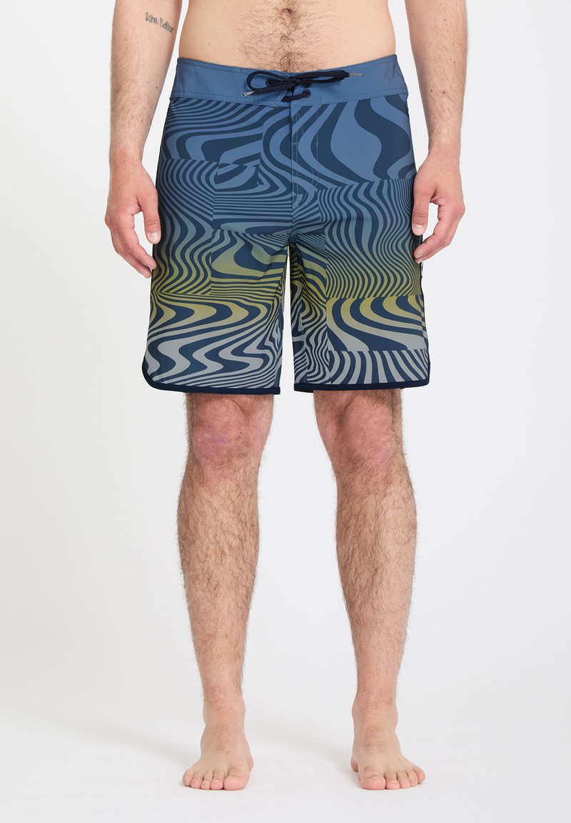 Homme portant un short de bain à motifs ondulés bleus et jaunes, debout pieds nus devant un fond blanc uni.
