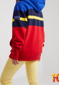 Sudadera con bloques de color en franjas azul, roja, marino y amarilla. Presenta un parche con logo en la manga y puños acanalados. Textura de tejido suave.