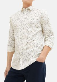Chemise blanche à boutons avec un motif de rayures verticales bleues et noires, manches longues retroussées aux poignets, et une poche poitrine. Texture de tissu lisse.