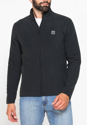 Uomo che indossa una giacca in pile nera con zip e piccolo logo sul petto e jeans azzurri, in piedi davanti a uno sfondo bianco.