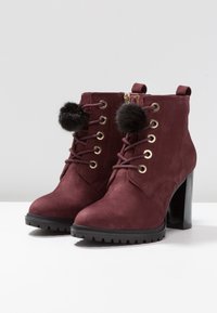 Botas de tobillo de gamuza burdeos con pompones negros, ojales dorados, diseño con cordones y un tacón alto y grueso. Suela de goma texturizada para un mejor agarre.